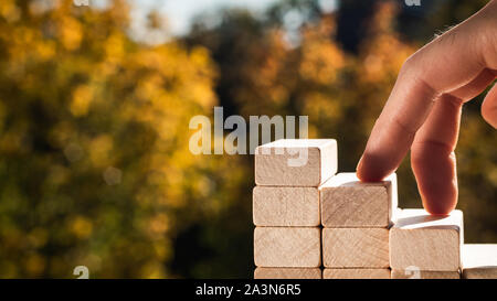 Concept de promotion de l'entreprise objectifs. Les doigts de l'homme monter les escaliers faits de blocs de bois contre l'arrière-plan de feuilles d'automne sur une journée ensoleillée. Banque D'Images