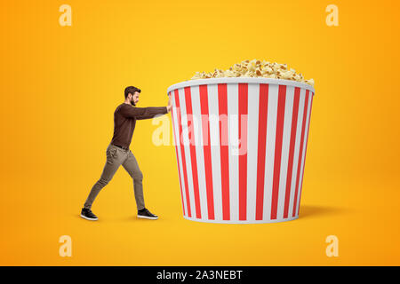 Vue latérale du jeune homme en oufit poussant d'énormes seau de pop corn sur fond orange. Banque D'Images