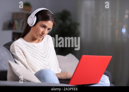 Jeune fille sérieuse portant des écouteurs contrôle de contenu portable rouge assis sur un canapé dans la nuit à la maison Banque D'Images