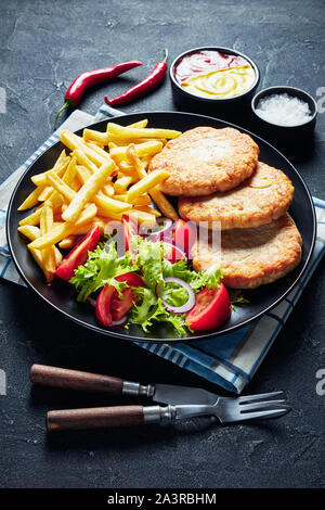 Close-up of fried turquie des hamburgers servis avec salade de tomate salade et frites sur une plaque noire sur une table en béton, avec du ketchup et de la moutarde, ve Banque D'Images