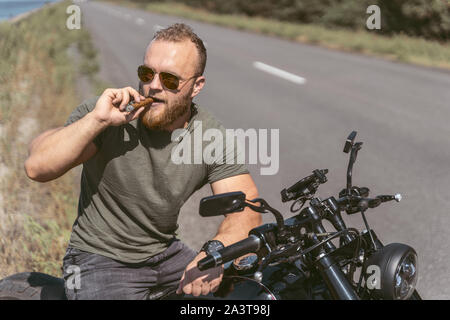 Brutal attrayant biker assis sur le vélo de sigar fumeurs sur le côté de la route Banque D'Images