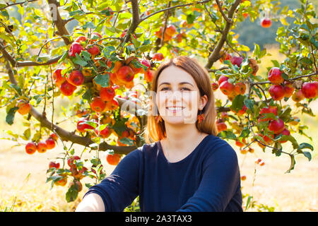 Young smiling woman enjoying in a sunny apple tree garden. Porte de fille fait main boucles d'élégant. Banque D'Images
