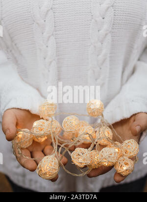 Lumières dans les paumes. Les mains tiennent une guirlande avec boules rondes en osier. Jeune fille dans un chandail blanc avec des lumières de Noël dans ses mains chaudes, Nouvelle Année Banque D'Images