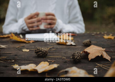Jeune femme dans un pull blanc est assis à une vieille table sombre dans le parc, est titulaire d'une tasse de thé avec du café et lit et écrit sur l'arrière-plan de jaune Banque D'Images