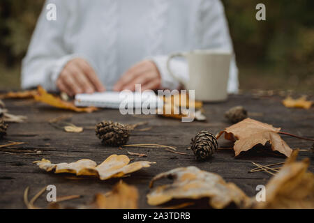 Jeune femme dans un pull blanc est assis à une vieille table sombre dans le parc, est titulaire d'une tasse de thé avec du café et lit et écrit sur l'arrière-plan de jaune Banque D'Images