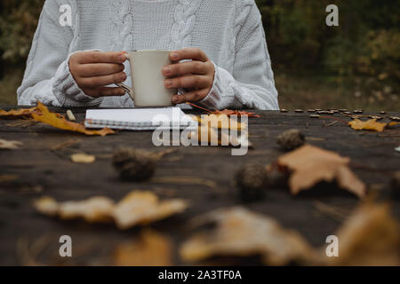 Jeune femme dans un pull blanc est assis à une vieille table sombre dans le parc, est titulaire d'une tasse de thé avec du café et lit et écrit sur l'arrière-plan de jaune Banque D'Images