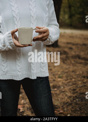 Jeune femme dans un pull blanc est titulaire d'une tasse de thé avec du café sur un fond de feuilles d'automne jaune. Automne chaud atmosphère, soft focus. Copy space Banque D'Images