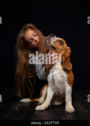 Une jeune fille rousse de 10 ans joue avec un chiot beagle assis sur un parquet en bois sombre en face d'un fond noir. Banque D'Images