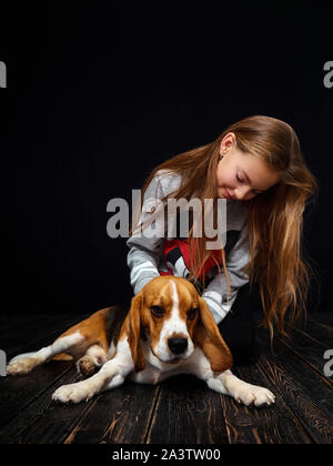 Une jeune fille rousse de 10 ans joue avec un chiot beagle assis sur un parquet en bois sombre en face d'un fond noir. Banque D'Images