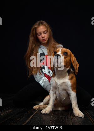 Une jeune fille rousse de 10 ans joue avec un chiot beagle assis sur un parquet en bois sombre en face d'un fond noir. Banque D'Images