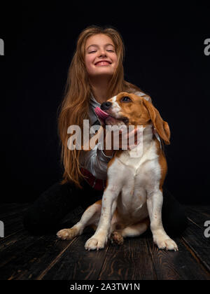 Une jeune fille rousse de 10 ans joue avec un chiot beagle assis sur un parquet en bois sombre en face d'un fond noir. Banque D'Images