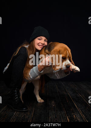 Une jeune fille rousse de 10 ans joue avec un chiot beagle assis sur un parquet en bois sombre en face d'un fond noir. Banque D'Images