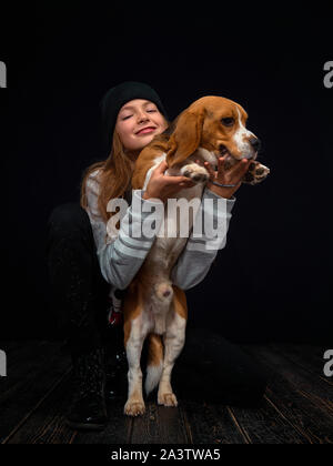 Une jeune fille rousse de 10 ans joue avec un chiot beagle assis sur un parquet en bois sombre en face d'un fond noir. Banque D'Images