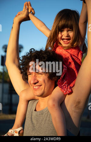 Jeune père avec sa petite fille sur ses épaules au coucher du soleil. L'homme est à l'écart d'élever ses mains, et elle est en train de rire avec la bouche ouverte l Banque D'Images