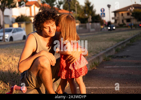 Peu de tout-petit fille sur un vélo à l'embrasser jeune père piscine au coucher du soleil. Copie de l'espace sur la droite. Banque D'Images