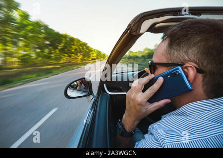 Businessman talking on cellphone en conduisant une voiture, vue de côté. Beau jeune homme en shirt talking on mobile phone tout en maintenant le lor Banque D'Images