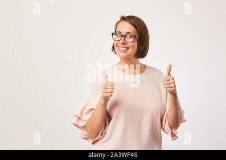 Portrait de la belle jeune femme en chemisier élégant portant lunettes et permanent showing Thumbs up over white background Banque D'Images