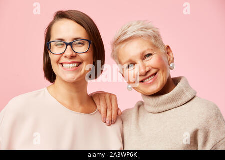 Portrait de belle fille à lunettes se tenant ensemble avec sa mère et ils smiling at camera sur fond rose Banque D'Images