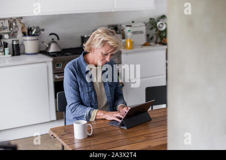 Adulte mature woman in kitchen en utilisant une tablette numérique avec clavier Banque D'Images