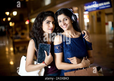 Deux jeunes femmes élégantes s'amusant avec tablette numérique et les écouteurs on city street at night Banque D'Images