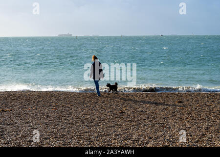 Une femme d'âge moyen promener son chien noir le long d'une plage de galets avec la mer en arrière-plan Banque D'Images
