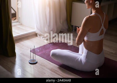 Femme méditant à l'intérieur. Attrayant et short-haired young model in white sportclothes assis sur le tapis et pratiquer le yoga avec des bougies. Priez Banque D'Images