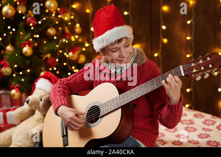 Teen boy à jouer de la guitare, assis près de l'arbre de Noël décoré avec des lumières, vêtu comme Santa helper - Joyeux Noël et bonnes fêtes ! Banque D'Images