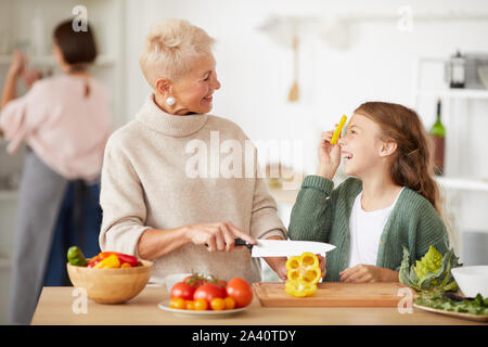 Petite fille s'amuser tandis que sa grand-mère poivre frais de coupe sur une planche à découper ils souriant l'un à l'autre tout en se tenant dans la cuisine Banque D'Images