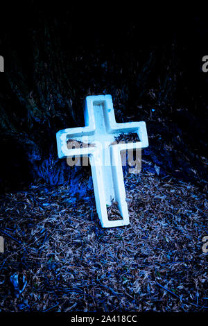 Une pierre tombale en forme de croix blanche entourée de feuilles tombées dans un ancien cimetière sombre Banque D'Images