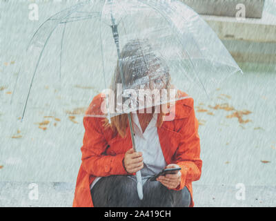 Caucasian girl avec gabardine rouge promenades dans un parc sous un parapluie transparent. Banque D'Images