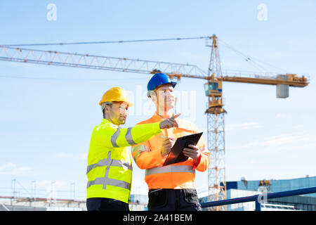 Deux jeunes ingénieurs masculins debout en face de la grue mécanique travaillant sur le chantier de construction Banque D'Images
