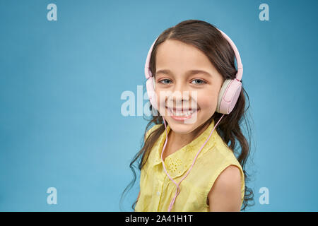 Funny Girl wearing headphones listening music, looking at camera and smiling in studio. Heureux l'enfant à l'aide d'écouteurs et profiter de chansons. Jolie teen femelle posant sur fond isolé bleu. Banque D'Images