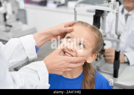 Vue du côté de belle femme contrôle d'oculiste eye élève de smiling girl en laboratoire. Cheerful patient testing vision dans une clinique et le choix des lunettes ou lentilles. Concept de l'examen. Banque D'Images