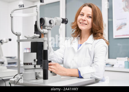 Belle femme contrôle de la vue avec l'oculiste à la lampe à fente en clinique. Yeux professionnel Médecin en uniforme blanc assis à table, looking at camera and smiling in magasin d'optique. Concept de soins des yeux. Banque D'Images