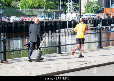 Un homme qui court, et un homme d'âge moyen avec une valise à roulettes à pied le long des quais à Newcastle Upon Tyne, Tyne and Wear, England, UK Banque D'Images