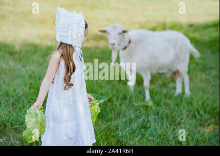 Un berger jeune fille en robe blanche et bonnet alimente une chèvre avec des feuilles de chou. L'alimentation de l'enfant au printemps chèvre domaine. Banque D'Images