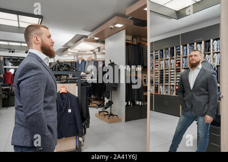 Vue latérale d'un jeune homme portant nouvelle veste, gardant la main dans la main, à la recherche de miroir et estimer ensemble en boutique. En choisissant des vêtements masculins graves shop. Concept de montage et l'élégance. Banque D'Images