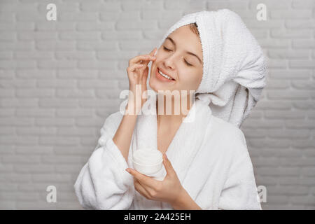 Jeune, jolie femme en blanc serviette avec la peau parfaite l'application sur le visage naturel crème pour hydrater et rajeunir. Concept de produit pour les soins de la peau du visage pour femme. Banque D'Images