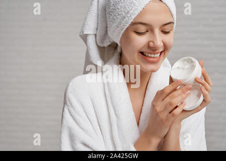 Belle, smiling model holding et montrant fort avec la crème hydratante pour le visage. Jeune femme à la santé, une peau parfaite en blanc peignoir avec une serviette sur la tête posant sur fond gris. Banque D'Images