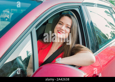 Happy smiling woman dans une voiture. La conduite. Portrait of young caucasian business dame en rouge vêtements looking at camera, assis derrière le volant rouge c Banque D'Images