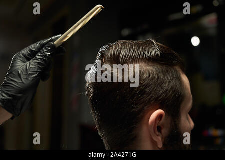 Close up of barbu avec tête humide se coupe par coiffure tout en restant assis dans un fauteuil au salon de coiffure. Coiffure en gants noirs se passe à peigner les cheveux et faire de nouveau style pour son client exigeant Banque D'Images