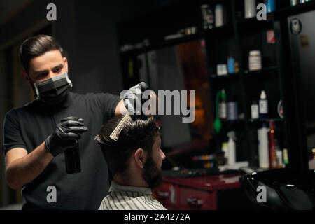 Beau barbu assis dans la chaise et couverts par cape rayée à barbershop coiffure hommes en noir chemise semble très concentré pendant le processus de travail. Traitement de beauté masculine Banque D'Images