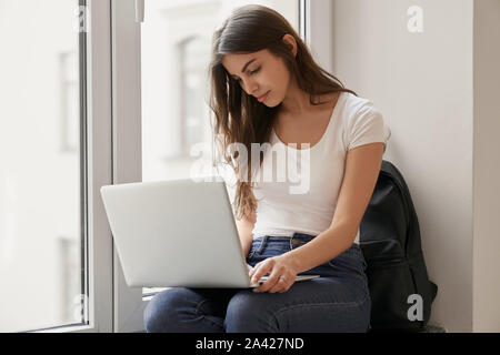 Portrait de jeune fille belle brunette en jupe blanche et bleu jeans assis sur la fenêtre et à la recherche à l'ordinateur qui est couchée sur ses genoux. La préparation de l'étudiant est exposé dans le hall de l'université Banque D'Images