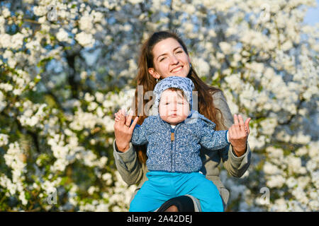 Tout-petit avec maman sur le fond d'arbres en fleurs. petit bébé sur les mains de la mère. femme jouant avec des enfants à l'extérieur dans le jardin de printemps en fleurs Banque D'Images