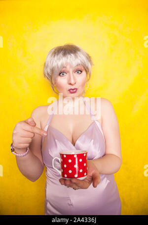 Jolie jeune femme blonde en courbes, avec les cheveux courts jouit de boire du thé à partir de la jolie tasse en céramique rouge à pois jaune sur fond de studio. Banque D'Images