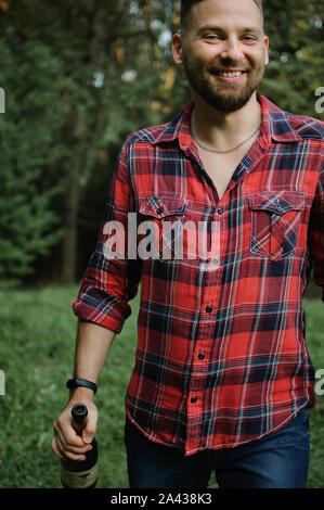 Hipster barbu en chemise rouge et bleu foncé jeans est hoolding une bouteille de vin et regardant la caméra avec sourire. La dépendance à l'alcool. Banque D'Images