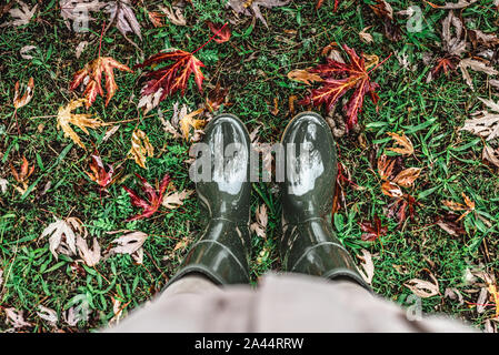 Les pieds dans des bottes en caoutchouc vert olive debout sur l'herbe verte avec des feuilles mortes. Banque D'Images