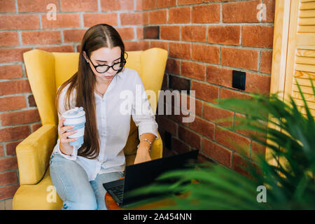 Jeune femme d'affaires réussie dans les verres de travailler sur un ordinateur portable noir dans un coffee shop, holding Coffee cup et la saisie. Banque D'Images