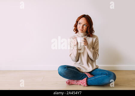 Jeune femme aux cheveux rouge rêve assis sur le plancher sur un arrière-plan blanc vide. Souriant modèle féminin à haut, tenant une tasse de café chaud. Banque D'Images