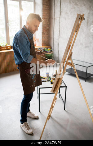 Homme en tablier, debout devant un chevalet et la peinture avec des couleurs de l'eau Banque D'Images